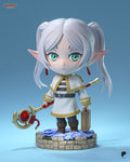 Sculptor's render of the painted design (kit ships unpainted): front view of the chibi elf mage with silver twin tails, holding a red gem staff and a small suitcase, on a round stone bed of blue flowers